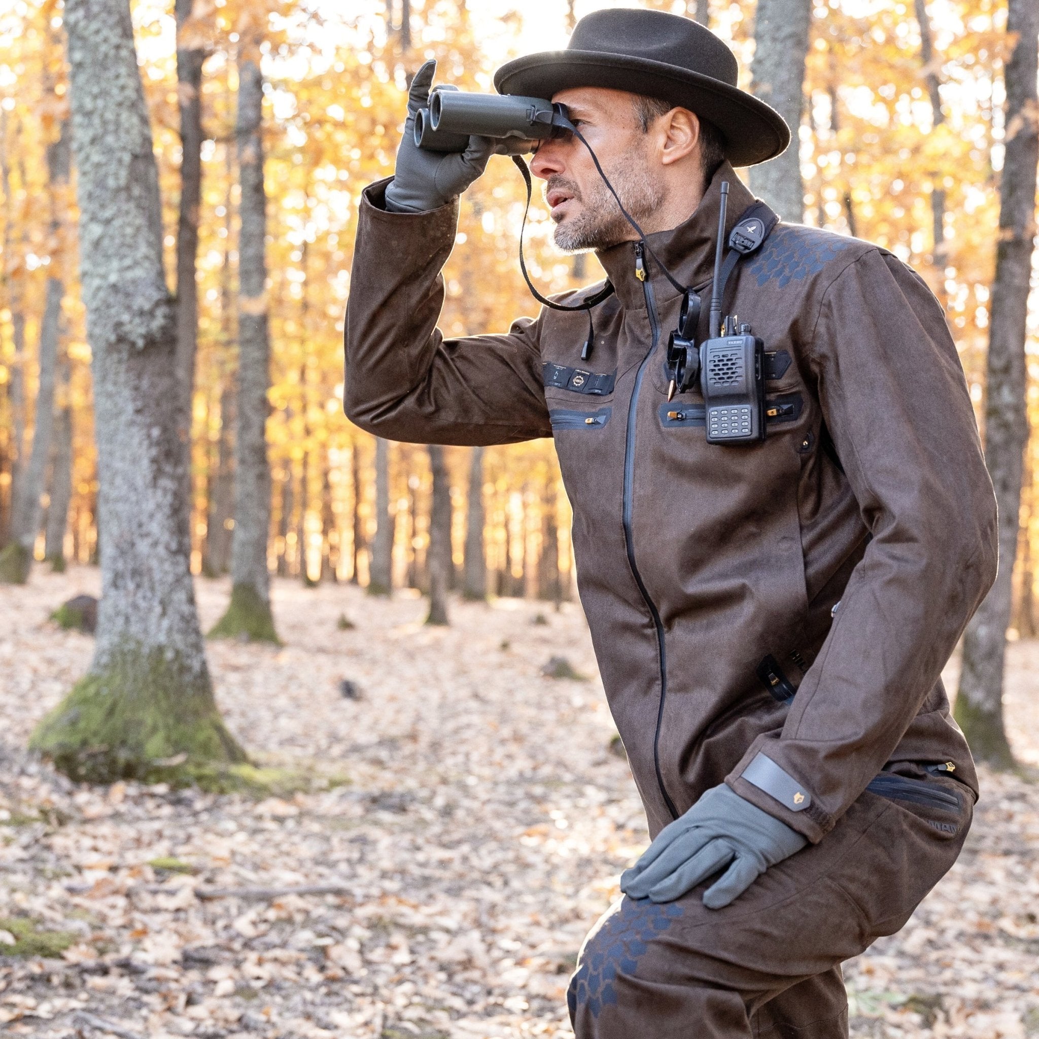 A person wearing a HILLMAN GEAR 6OL Cold Weather Hunting Jacket, gloves, and a hat is kneeling among trees with autumn leaves, looking through binoculars. A radio is clipped to the jacket.