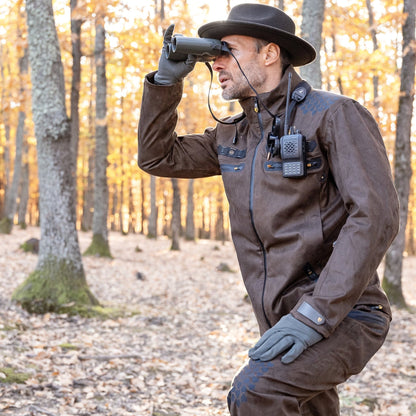 A person wearing a HILLMAN GEAR 6OL Cold Weather Hunting Jacket, gloves, and a hat is kneeling among trees with autumn leaves, looking through binoculars. A radio is clipped to the jacket.