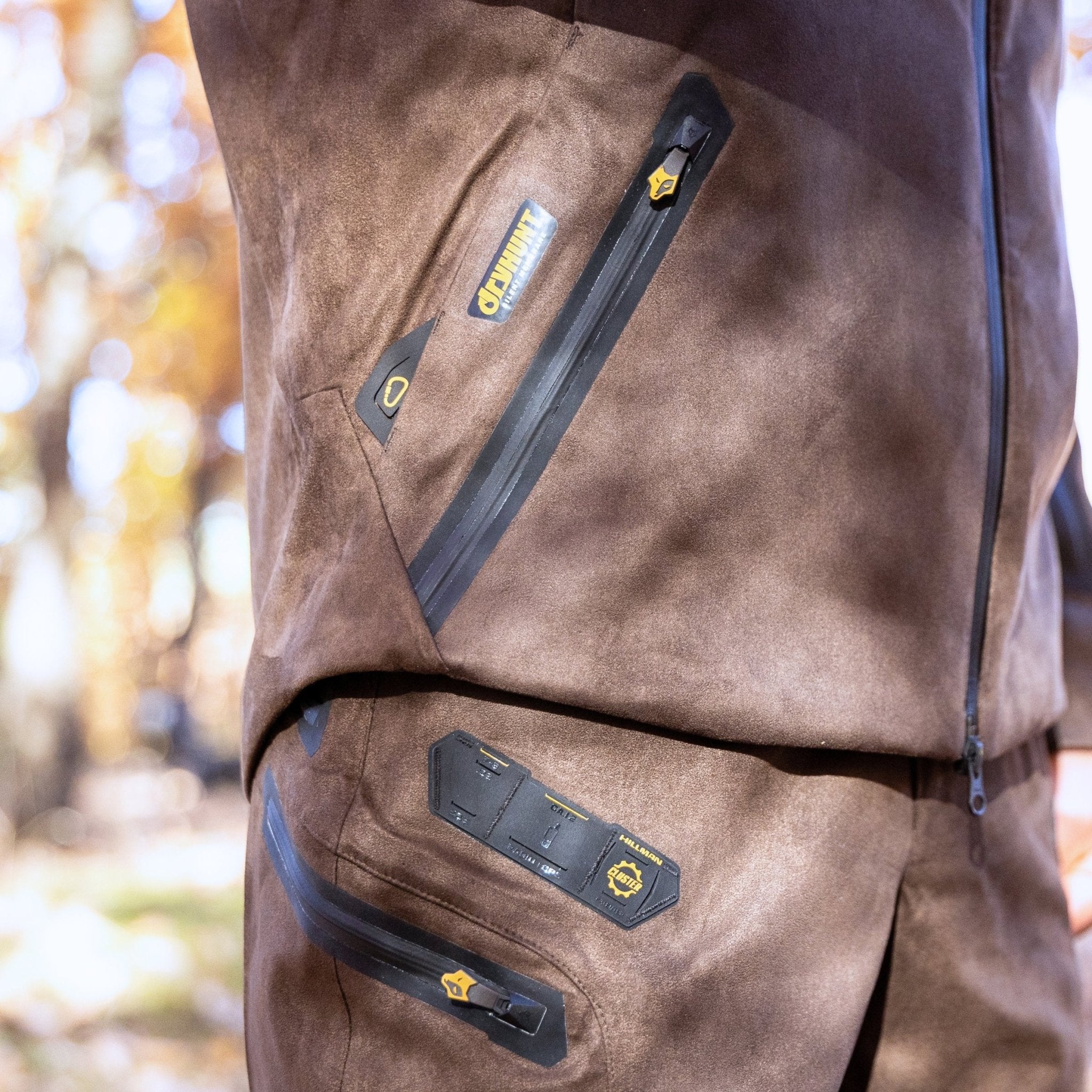 A close-up of a person wearing the HILLMAN GEAR Camo Hunting Jacket 6OL Heated - Cold Weather Insulated, featuring black zippers and yellow logo tags. The background has blurred autumn foliage in an outdoor setting.