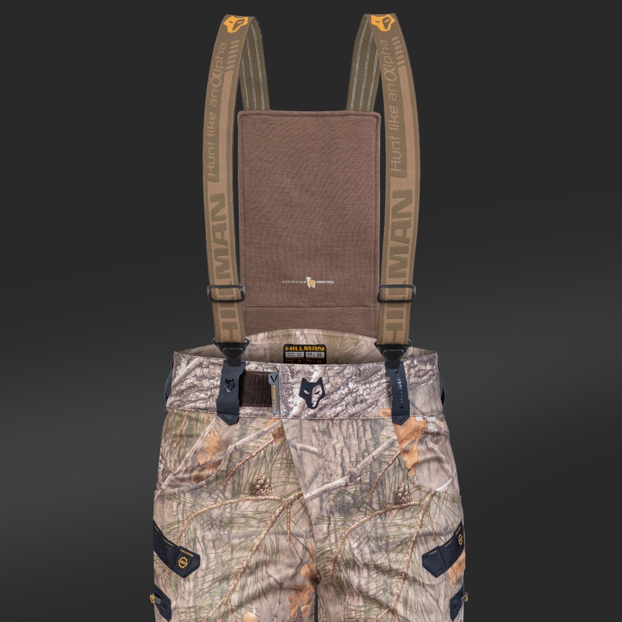 The 6OL Insulated Camo Hunting Pants by HILLMAN GEAR feature waterproof camouflage bibs with adjustable HUMAN labeled suspenders, buckles, multiple pockets, and an animal graphic logo on the waistband. They offer extreme cold protection against a solid dark gray background.