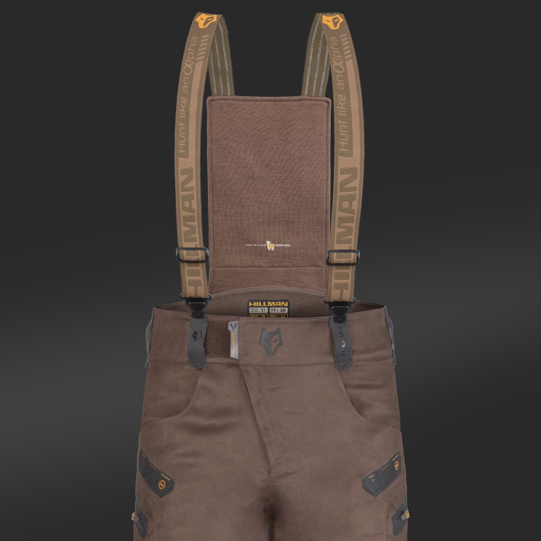 Brown suspenders connect rugged, insulated 6OL Matrix Hunting Pants by HILLMAN GEAR. These pants have black patches, reinforced edges, waterproof bibs with multiple pockets and belt loops, all set against a dark backdrop. The HILLMAN name is prominent on the suspenders and waistband.