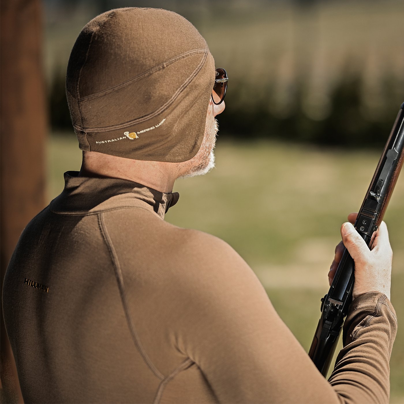 Wearing a Hillman® Anatomic Merino Beanie and a brown thermo-regulating merino wool jacket, an individual, their features obscured by sunglasses and a hat, holds a rifle amidst blurred green grass and wooden post backdrop.