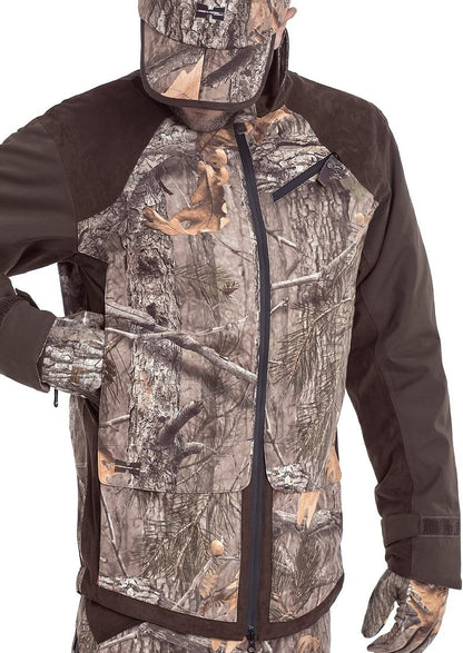 A person wears a Hillman® Fusion Hunting Jacket 3DX with ultra-light hollow fiber insulation. The camouflage pattern of tree trunks and leaves blends into the forest, while zippers and black fabric accents enhance functionality. They also wear matching gloves and hat.