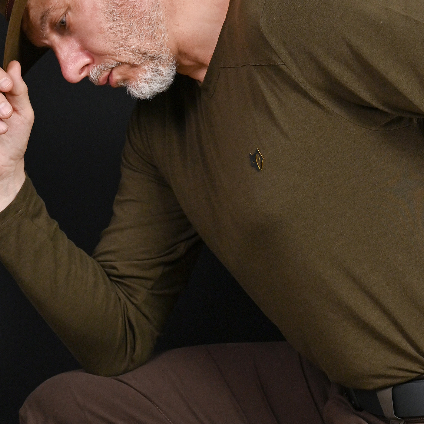 A man in a HILLMAN 1UL Bamboo Long Sleeve Hunting T-Shirt rests his elbow on his knee, clutching a brown hat, with moisture control features helping him stay dry. He wears brown pants as shadows partially obscure his bearded face against the dark background.