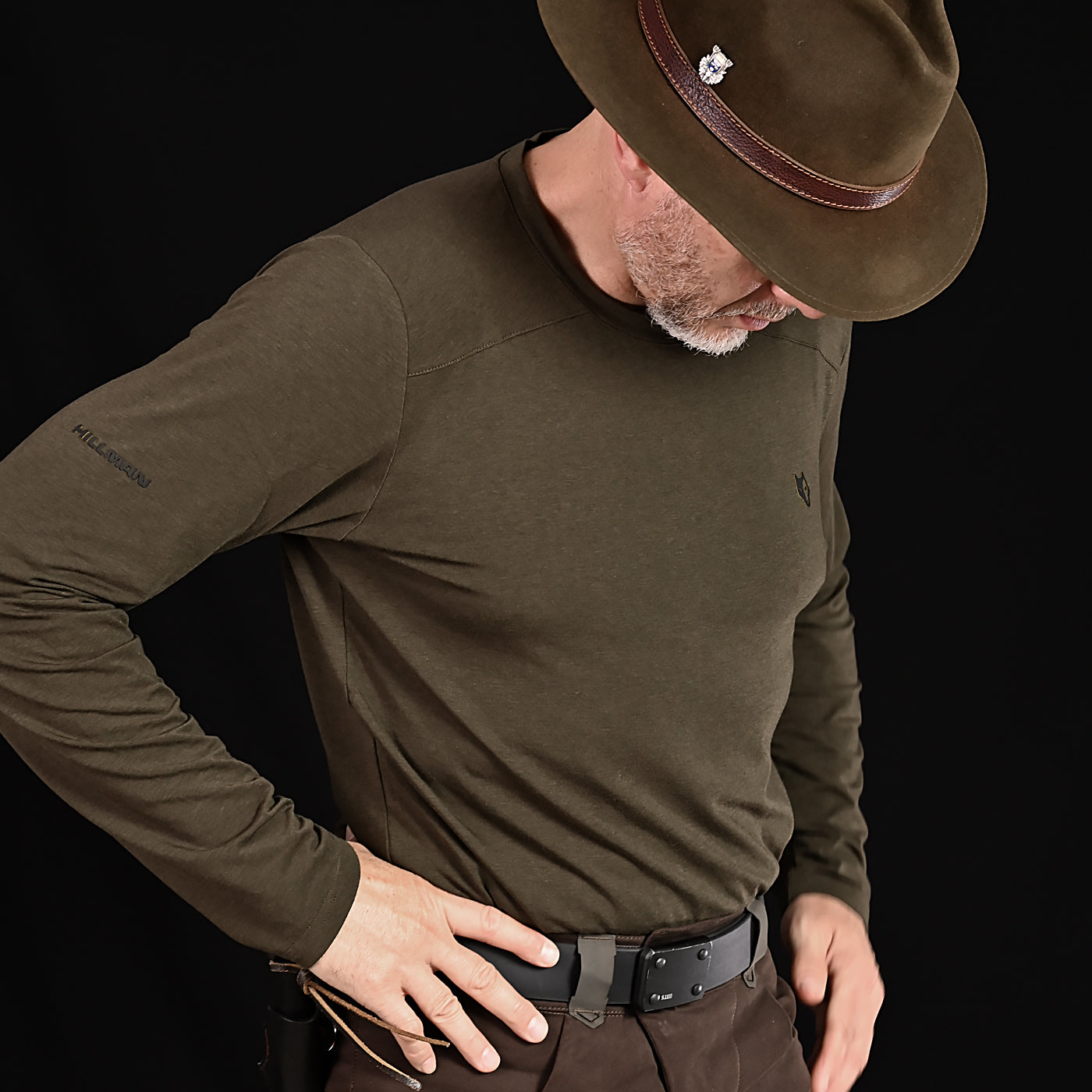 A person stands against a black background wearing the HILLMAN 1UL Bamboo Long Sleeve Hunting T-Shirt, an ultralight all-season base layer. Crafted for moisture control and paired with a brown hat and pants, they look down with hands near their belt.